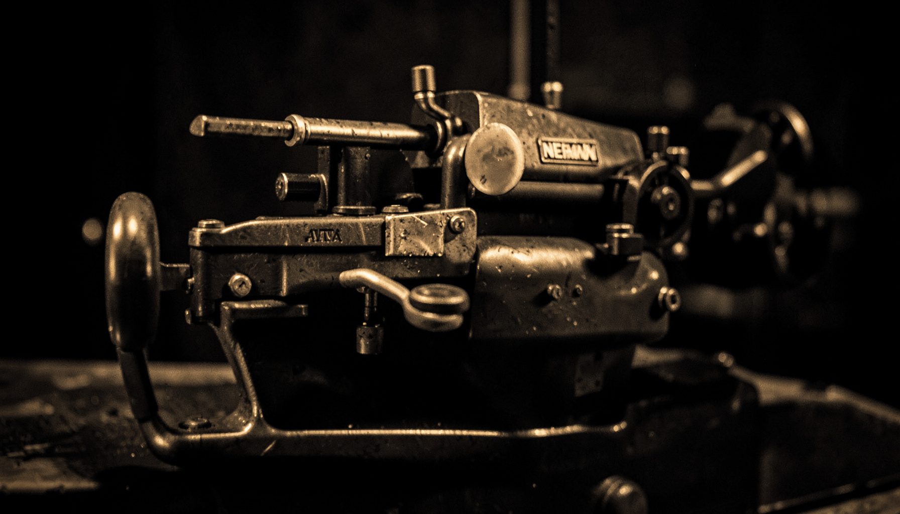 A warm duotone photograph of a vintage Neumann lathe in a dimly lit studio, its metallic surfaces catching amber light against deep charcoal shadows.